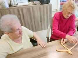 Zwei ältere Frauen sitzen an einem Tisch in einem hellen Raum. Eine Frau mit grauen Haaren lächelt, während die andere in einem pinken Pullover mit einem langen Teigstück arbeitet. Um sie herum sind verschiedene Alltagsgegenstände zu sehen.