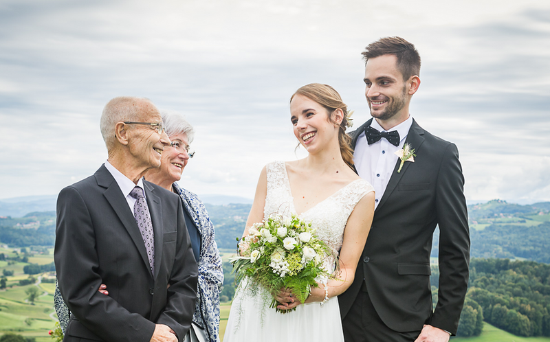 Hochzeitsfeier mit einem Brautpaar und zwei älteren Gästen im Freien. Die Braut trägt ein elegantes weißes Kleid und hält einen Blumenstrauß. Der Bräutigam ist in einem schwarzen Anzug. Im Hintergrund sind grüne Hügel und ein bewölkter Himmel zu sehen.