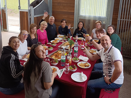 Eine Gruppe von 12 Personen sitzt an einem Tisch mit rotem Tischdecken und genießt ein gemeinsames Essen. Es sind Teller mit Essen und Getränke auf dem Tisch. Einige Personen winken und lächeln in die Kamera. Im Hintergrund sind Fenster und Holzstrukturen sichtbar.