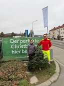 Zwei Personen stehen neben einem Weihnachtsbaum und einem Werbeschild.