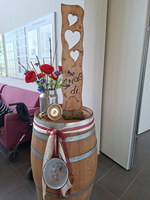 Ein dekoratives Holzschild mit Herzen steht auf einem Holzfass, das mit einem roten und weißen Band geschmückt ist. In der Mitte befindet sich ein silberner Behälter mit bunten Blumen. Im Hintergrund ist ein Teil eines Sitzbereichs sichtbar.