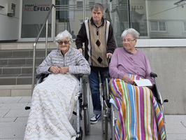 Drei Personen im Rollstuhl vor einem Gebäude, eine Person steht hinter ihnen.