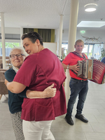 Pflegekraft umarmt Seniorin, während ein Musiker im Hintergrund Akkordeon spielt.