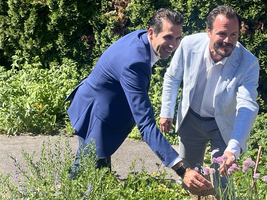 Zwei Männer stehen in einem Garten und beugen sich über eine Gruppe von Pflanzen. Der Mann links trägt einen blauen Anzug, während der Mann rechts in einem hellen Anzug gekleidet ist. Beide lächeln und scheinen sich an den Pflanzen zu erfreuen, umgeben von grünen Sträuchern.