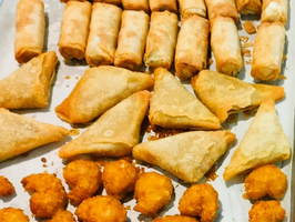 Ein Teller mit verschiedenen frittierten Snacks, darunter Frühlingsrollen, Teigtaschen und frittierte Kugeln. Die Snacks sind goldbraun und auf einer weißen Unterlage angeordnet.