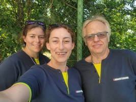 Drei Personen lächeln in blauen Shirts vor einem grünen Hintergrund.
