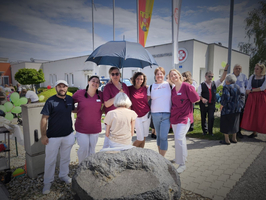 Eine Gruppe von sechs Personen in Arbeitskleidung steht lächelnd vor einem Gebäude. Einige Menschen sind im Hintergrund zu sehen, während eine Person mit einem Regenschirm im Vordergrund sitzt. Es herrscht eine festliche Atmosphäre im Freien.