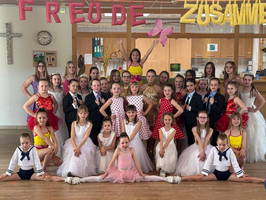 Eine Gruppe von Kindern und Jugendlichen steht in einem Raum, umgeben von bunten Dekorationen mit den Worten „Freude“ und „Zusammen“. Sie sind in verschiedenen Kostümen gekleidet, einige tragen Tanzkleidung, andere formelle Kleidung, und posieren gemeinsam für ein Foto.