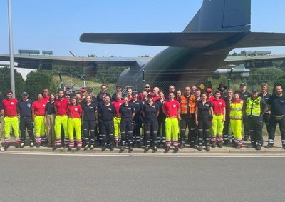 Eine Gruppe von etwa 40 Personen steht vor einem großen Flugzeug. Sie tragen helle Arbeitskleidung in Neonfarben und verschiedene Uniformen. Die Versammlung scheint aus Rettungs- und Einsatzkräften zu bestehen, die sich zur Kamera gruppiert haben, während sie in einem sonnigen, klaren Umfeld stehen.