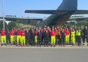 Eine Gruppe von etwa 40 Personen steht vor einem großen Flugzeug. Sie tragen helle Arbeitskleidung in Neonfarben und verschiedene Uniformen. Die Versammlung scheint aus Rettungs- und Einsatzkräften zu bestehen, die sich zur Kamera gruppiert haben, während sie in einem sonnigen, klaren Umfeld stehen.
