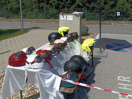 Eine Reihe von Motorradhelmen in verschiedenen Farben steht auf einem Tisch, der mit einem weißen Tuch bedeckt ist. Neben den Helmen sind zwei gelbe Helme sichtbar. Ein Sonnenschirm schützt den Bereich vor direkter Sonneneinstrahlung. Im Hintergrund sind Bäume und ein Parkplatz sichtbar.