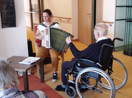 Eine Frau spielt Akkordeon, während ein älterer Mann im Rollstuhl zuhört.