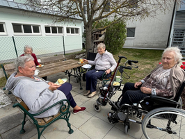 Vier ältere Frauen sitzen auf einer Terrasse, einige in Rollstühlen. Sie genießen Kuchen und Getränke, während sie sich unterhalten. Im Hintergrund steht ein Baum und ein Gebäude. Die Atmosphäre wirkt entspannt und gesellig.