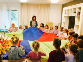 Eine Gruppe von etwa 20 Kindern und zwei Erwachsenen steht im Raum und hält gemeinsam ein großes, farbenfrohes Tuch. Die Kinder lächeln und scheinen aktiv am Spiel teilzunehmen, während eine ältere Frau und eine Frau mit einer Bluse in der Mitte stehen und das Spiel anleiten.