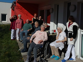 Eine Gruppe von neun älteren Menschen und zwei Pflegekräften steht vor einem Gebäude. Einige sitzen in Rollstühlen. Sie lächeln und scheinen sich zu amüsieren. Der Himmel ist blau mit wenigen Wolken. Die Wand des Gebäudes ist rot.