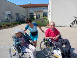 Eine Frau schiebt zwei ältere Menschen in Rollstühlen an einem sonnigen Tag.