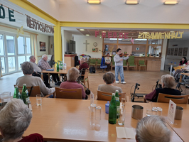 Ein Musiker steht im Vordergrund und spielt ein Instrument für eine Gruppe älterer Menschen, die an Tischen sitzen. Die Atmosphäre ist freundlich, mit bunten Bannern, die Freude und Gemeinschaft betonen. Im Hintergrund ist eine einladende Einrichtung zu sehen.