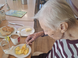 Eine ältere Frau sitzt an einem Tisch und isst Rührei von einem weißen Teller. Sie trägt ein gestreiftes Oberteil und hat graue Haare. Auf dem Tisch sind ein Glas Wasser, eine Tasse Kaffee, Butter und ein Brötchen zu sehen.