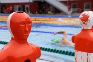 Zwei orangefarbene Übungsfiguren am Rand eines Schwimmbeckens, ein Schwimmer im Hintergrund.