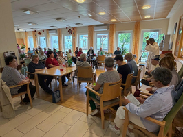 Eine Gruppe von etwa 20 Personen sitzt in einem hellen Raum mit großen Fenstern. Sie sind in einem Gespräch oder einer Aktivität vertieft. Einige Menschen halten Bücher oder Notizen. Tische und Stühle sind im Raum verteilt, und es gibt Pflanzen im Hintergrund.