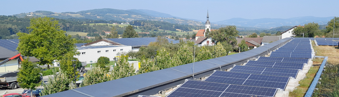 Dachinstallation der Photovoltaikanlage mit einer Aussicht auf eine Landschaft mit Hügeln, Gebäuden und einem Kirchturm im Hintergrund. Die Sonne scheint auf die Solarpanele.