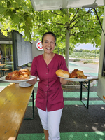 Eine Frau mit einem freundlichen Lächeln trägt auf zwei Tellern verschiedene Gerichte. Sie steht an einem Tisch im Freien, umgeben von grünen Bäumen und einem modernen Gebäude im Hintergrund. Die Szenerie vermittelt eine angenehme und einladende Atmosphäre.