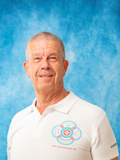 Ein älterer Mann lächelt vor einem blauen Hintergrund in einem weißen Poloshirt.