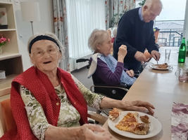 Drei ältere Menschen sitzen an einem Tisch in einem hellen Raum. Eine Frau mit einer roten Weste lächelt und hält einen Teller mit Essen. Eine andere Frau sitzt neben ihr, während ein Mann im Hintergrund sein Essen serviert.