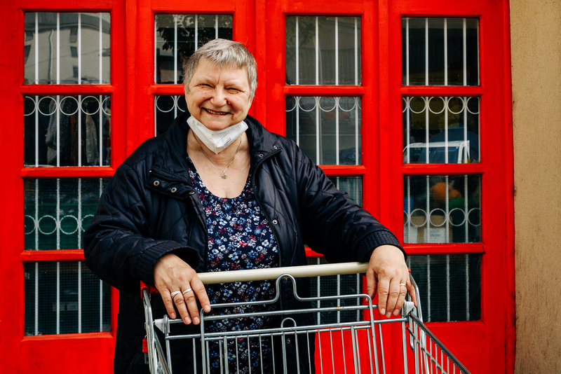 Eine ältere Dame, mit Maske unter dem Kinn und Einkaufswagen in der Hand, steht draußen vor einer roten Tür, Sozialmarkt, Böckhgasse