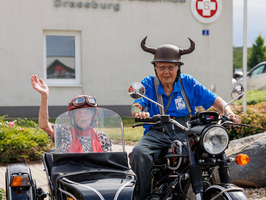 Zwei ältere Menschen auf einem Motorrad mit Beiwagen, fröhlich lächelnd.