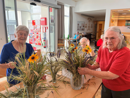 Drei ältere Frauen stehen an einem Tisch und gestalten Pflanzenarrangements mit frischen Blumen und Gräsern. In einem hellen Raum hinter ihnen sind weitere Personen sichtbar, die sich unterhalten oder beschäftigt sind.