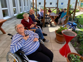 Ältere Menschen in Rollstühlen sitzen entspannt unter einem Sonnenschirm im Garten.