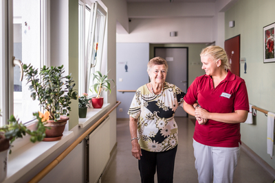 Eine ältere Dame wird freundlich von einer Pflegekraft in einem Flur begleitet.