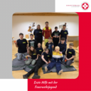Eine Gruppe von Jugendlichen in T-Shirts mit dem Aufdruck „Feuerwehrjugend“ posiert in einem Raum. Im Vordergrund sitzt eine Übungspuppe. Einige Jugendliche halten kleine Geräte. Im Hintergrund steht ein Erwachsene in Rettungsdienstuniform. Die Atmosphäre ist freundlich und lehrreich.