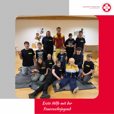 Eine Gruppe von Jugendlichen in T-Shirts mit dem Aufdruck „Feuerwehrjugend“ posiert in einem Raum. Im Vordergrund sitzt eine Übungspuppe. Einige Jugendliche halten kleine Geräte. Im Hintergrund steht ein Erwachsene in Rettungsdienstuniform. Die Atmosphäre ist freundlich und lehrreich.