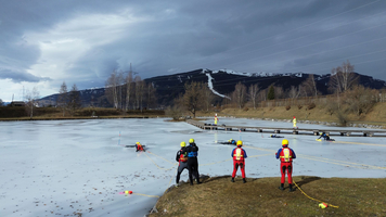 Rettungskräfte in speziellen Anzügen führen eine Übung auf einer zugefrorenen Fläche durch. Einige Teammitglieder sind im Wasser, während andere sicherheitsrelevante Aufgaben an Land übernehmen. Im Hintergrund sind schneebedeckte Berge und ein bewölkter Himmel sichtbar.