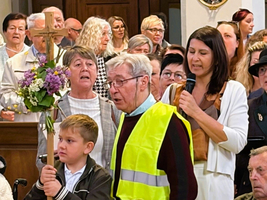 Eine Gruppe von Menschen in einer Kirche, die sich versammelt hat. Im Vordergrund stehen ein älterer Mann mit einer Weste, ein Junge mit einem Kreuz und zwei Frauen, die singen oder reden. Im Hintergrund sind viele weitere Personen zu sehen.