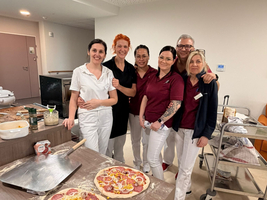 Sechs Personen in einheitlicher Kleidung stehen in einer Küche. Auf einem Tisch liegen zwei belegte Pizzen. Im Hintergrund sind Küchenutensilien und eine Rollwaage sichtbar. Die Gruppe scheint fröhlich und entspannt.