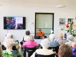 Ein Mann in orangefarbener Kleidung spricht vor einer Gruppe älterer Menschen, die in Rollstühlen sitzen. Im Hintergrund ist ein Bildschirm mit einem Bild von Motorradtaxis zu sehen. Die Atmosphäre wirkt freundlich und informativ.