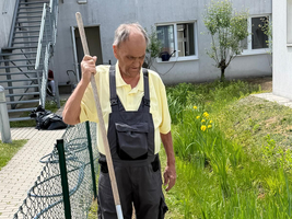 Ein älterer Mann in einem gelben T-Shirt und Latzhose steht im Garten und benutzt eine Harke, um den Boden zu bearbeiten. Im Hintergrund sind grüne Pflanzen und einige gelbe Blumen zu sehen.