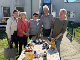 Fünf Menschen stehen an einem Tisch im Freien, der mit verschiedenen Gläsern gefüllt ist. Sie lächeln, während sie die Gläser betrachten. Die Umgebung ist hell und freundlich, mit einem Gebäude im Hintergrund.
