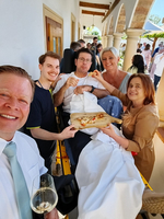 Eine Gruppe von fünf Personen steht vor einem Gebäude. Eine Person liegt auf einer Trage und hält eine Pizza. Die anderen vier Personen lächeln und zeigen auf die Pizza. Es ist ein sonniger Tag, und es gibt eine festliche Atmosphäre.