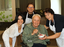 Eine Gruppe von vier Personen, darunter ein älterer Mann im Rollstuhl, der eine Schüssel mit Essen hält. Alle lächeln und tragen Schürzen. Die Umgebung wirkt freundlich und einladend, mit hellen Lichtverhältnissen und etwas Grüner Pflanzen.
