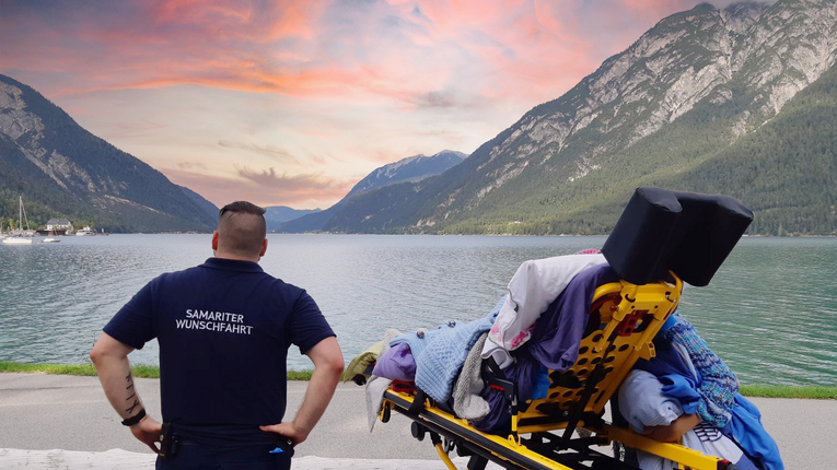 Wunschfahrt-Mitarbeiter mit Person auf Trage vor See und Bergen im Hintergrund